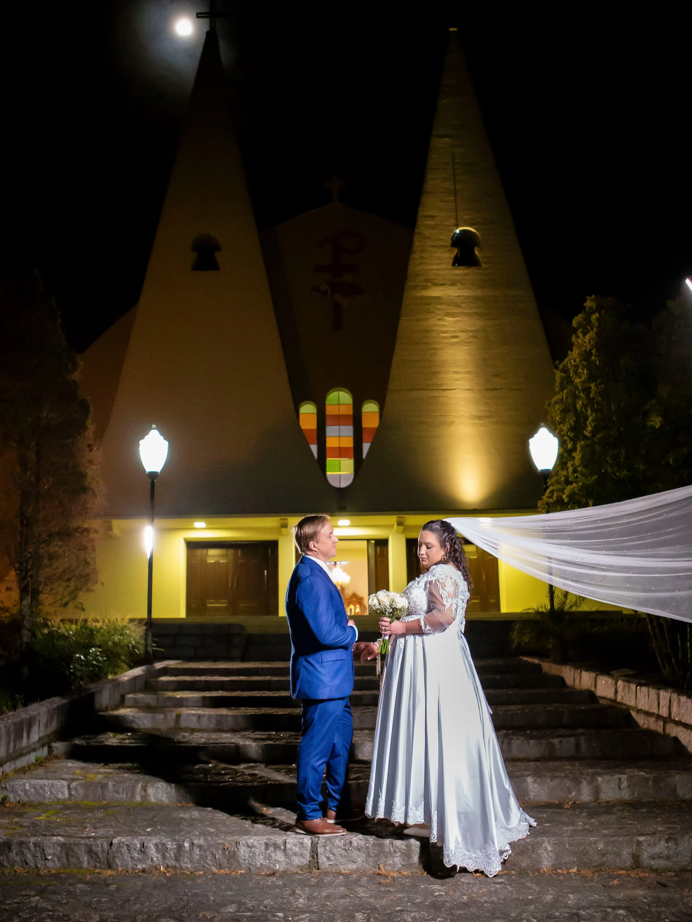 Noivos nos degraus de uma igreja iluminada à noite.