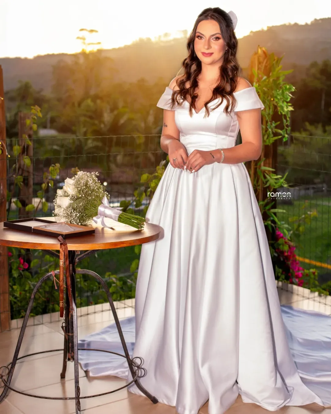 Noiva com buquê branco diante da paisagem ao pôr do sol.