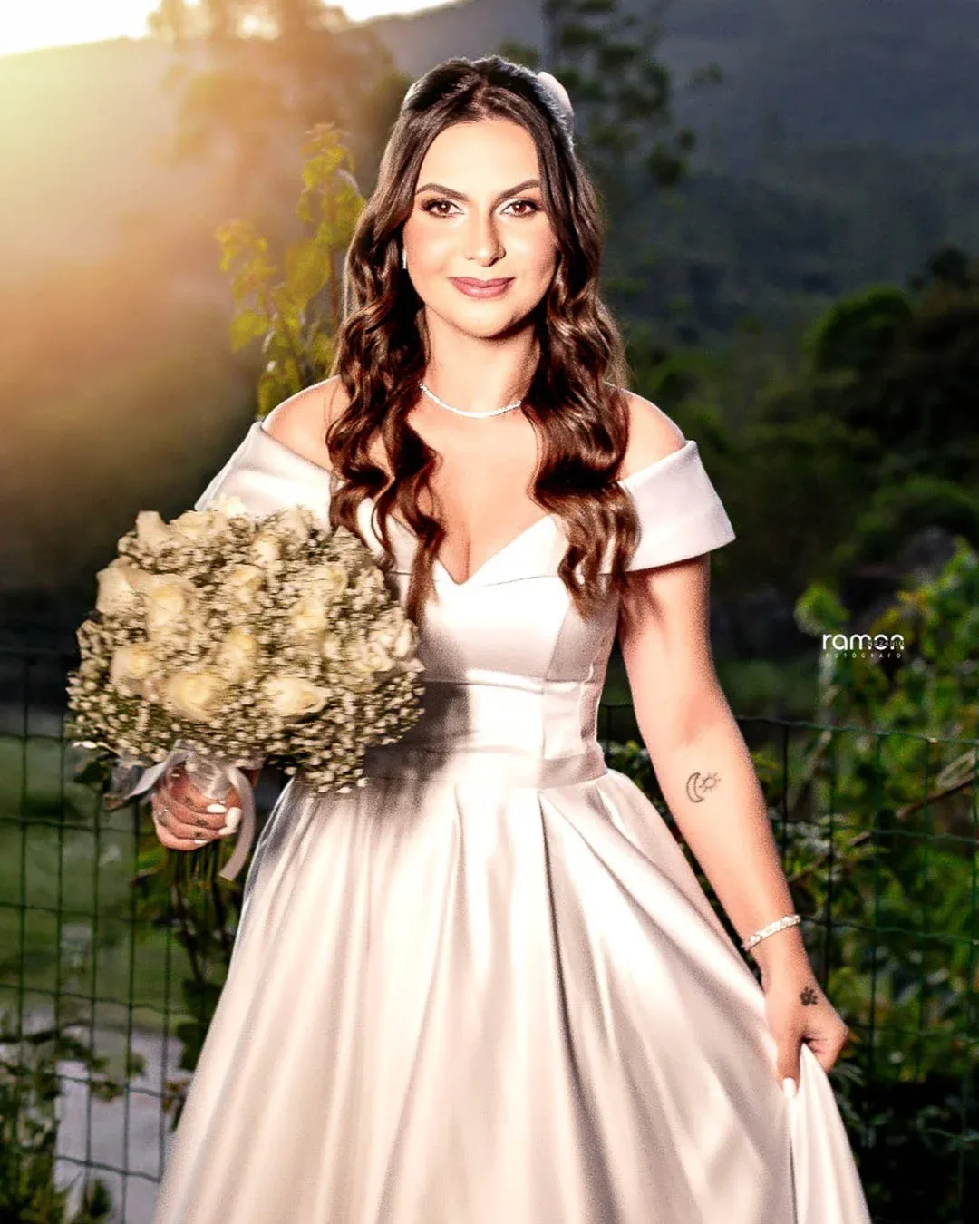 Noiva com vestido acetinado e buquê em uma paisagem ao fim da tarde.