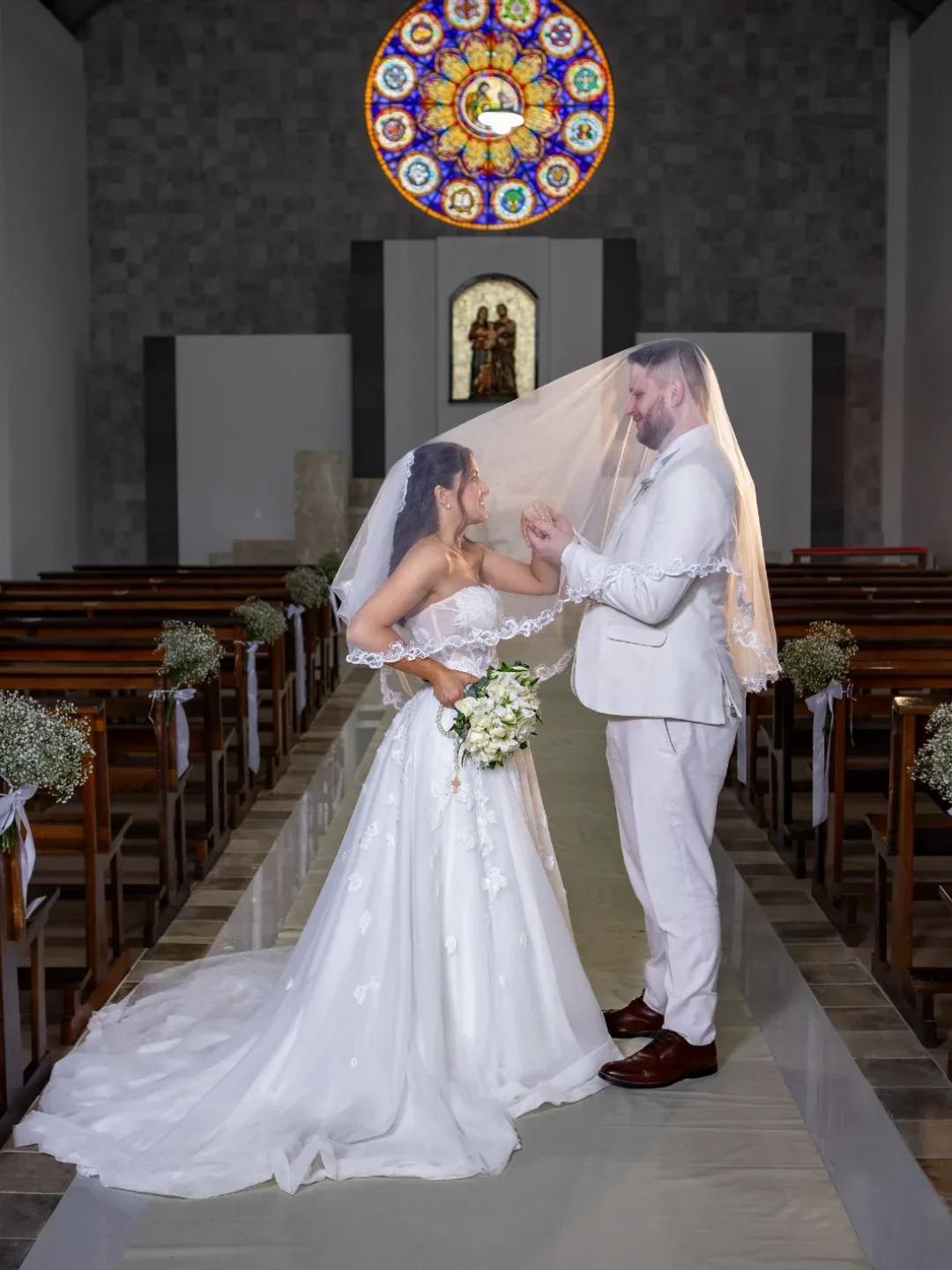 Noivos sob o véu diante do vitral de uma igreja.