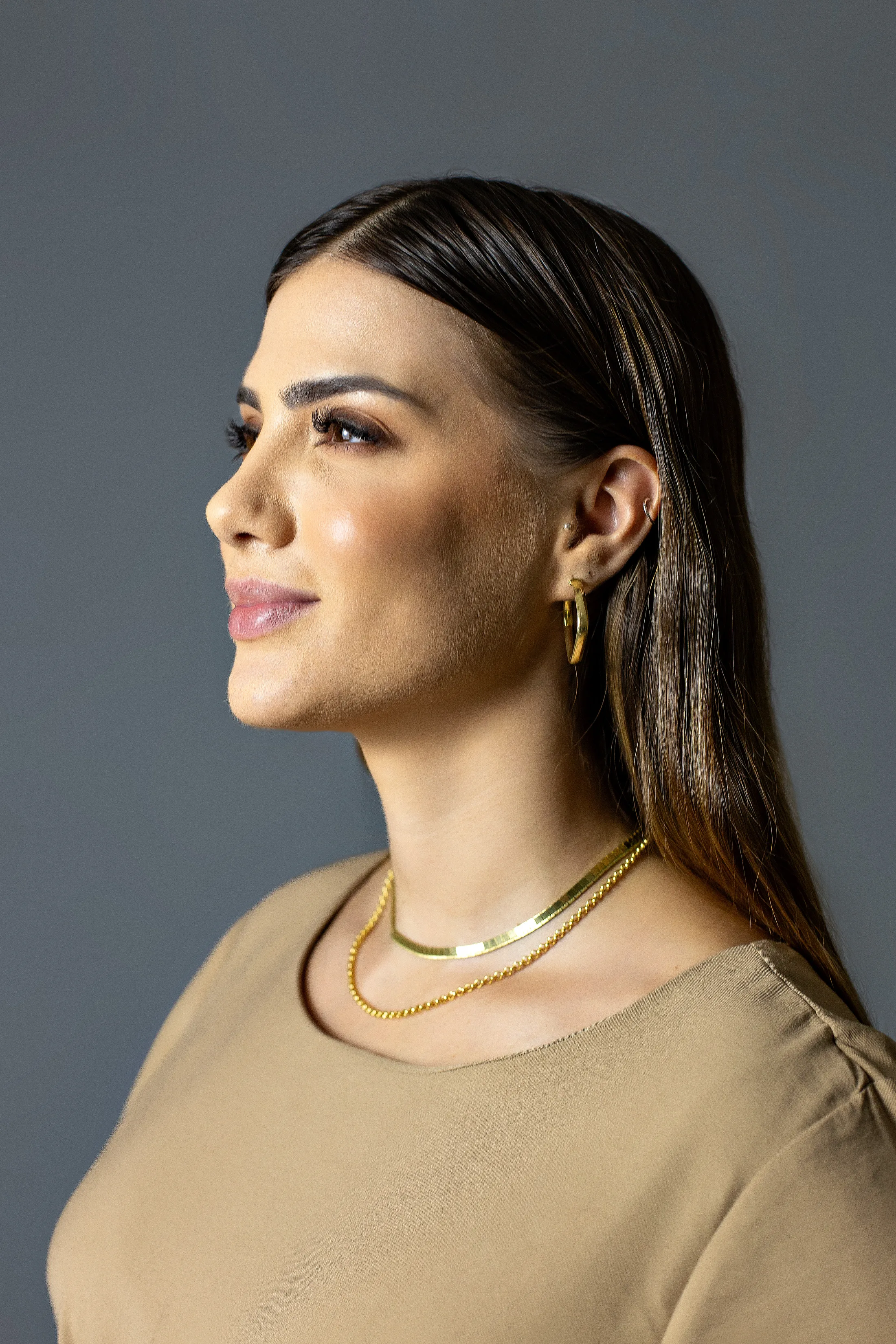 Retrato de uma mulher com blusa bege e colares dourados diante de fundo cinza.