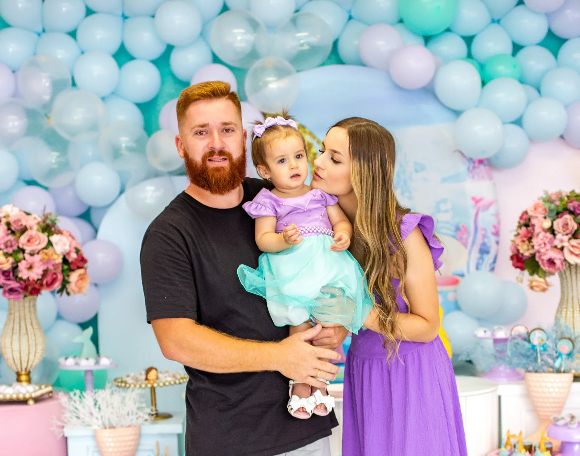 Dois adultos e uma criança pequena diante de balões coloridos em uma festa infantil.