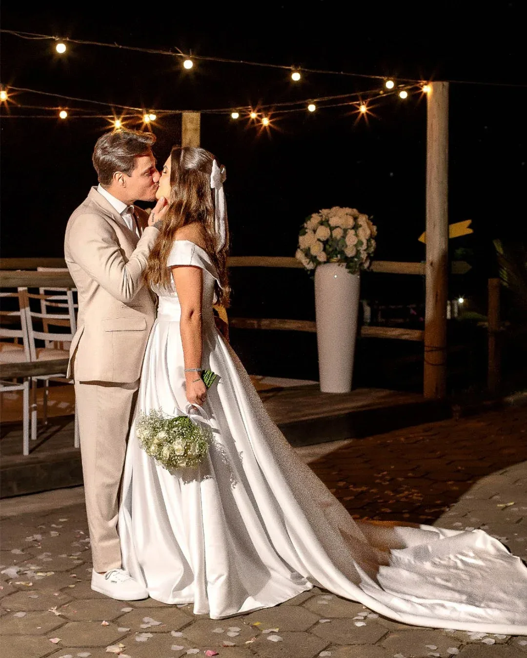 Noivos se beijando ao ar livre sob um varal de luzes.
